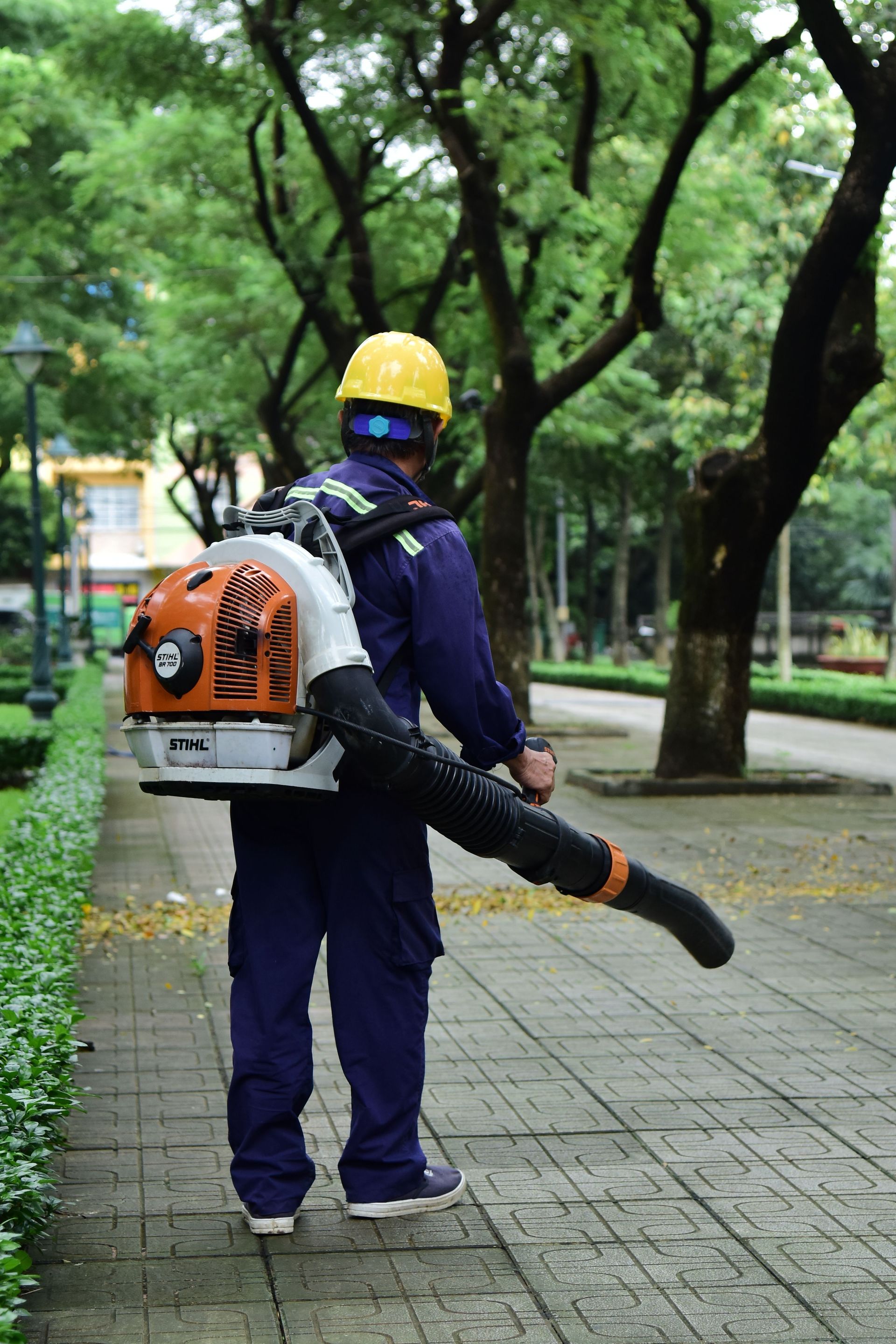 A city park worker in blue uniform and helmet operating a leaf blower on a walkway.